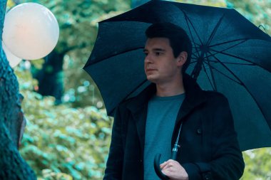 Yağmurlu günde şemsiyeli genç dalgın yakışıklı adam parkta yürüyüşe çıkıyor.