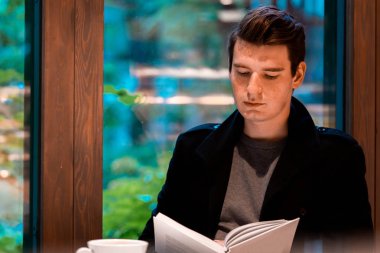 Genç dalgın yakışıklı adam kitap okuma ve rahat modern kafede yağmurlu bir günde çay veya kahve içme