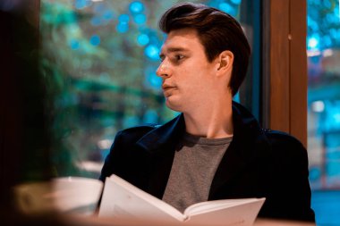 pensive adam kitap okuma ve rahat modern kafede yağmurlu bir günde çay veya kahve içme ve pencereden bakarak