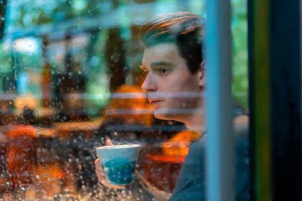 Young pensive handsome man drinking tea or coffee on rainy day in cozy modern cafe and looking through the window.