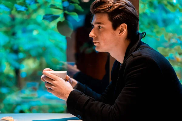 Jeune homme cher beau buvant du thé ou du café le jour de pluie et regardant par la fenêtre . — Photo