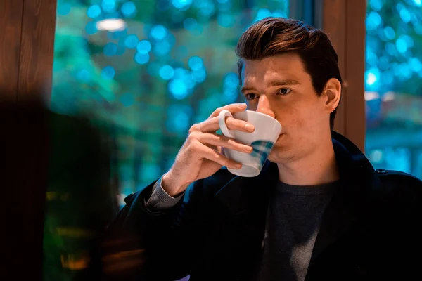 Jeune homme cher beau buvant du thé ou du café le jour de pluie et regardant par la fenêtre . — Photo