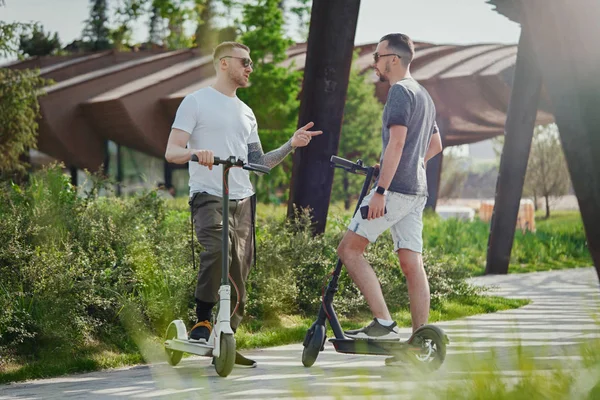 Zwei gutaussehende Männer unterhalten sich neben Elektro-Tretroller in Parklandschaft — Stockfoto