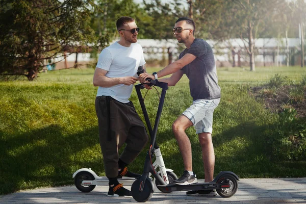 Zwei gutaussehende Männer unterhalten sich neben Elektro-Tretroller in Parklandschaft — Stockfoto