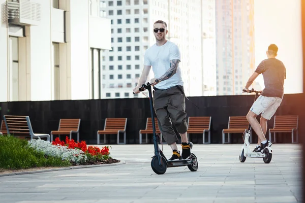 Dois homens bonitos montando scooters pontapé elétrico na bela paisagem urbana . — Fotografia de Stock