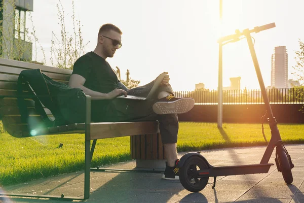 Hombre con portátil y scooter eléctrico moderno sentado en el parque contemporáneo . —  Fotos de Stock