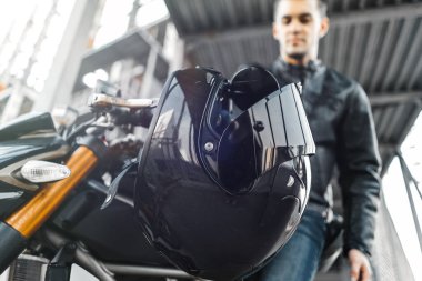 Portrait of handsome biker sitting on motorcycle next to black glossy helmet. clipart