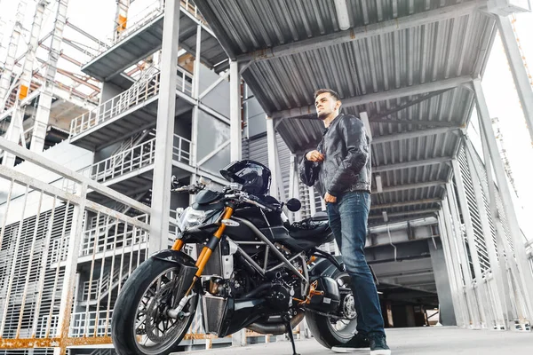 Retrato de motero guapo de pie junto a la motocicleta que se pone la chaqueta de cuero . —  Fotos de Stock