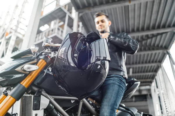 Retrato de motociclista bonito sentado na motocicleta ao lado de capacete preto brilhante . — Fotografia de Stock