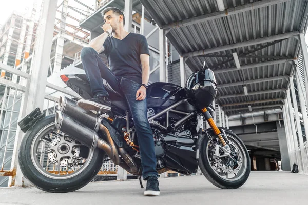 Motociclista guapo sentado en la motocicleta negra en el fondo urbano en el aparcamiento . —  Fotos de Stock