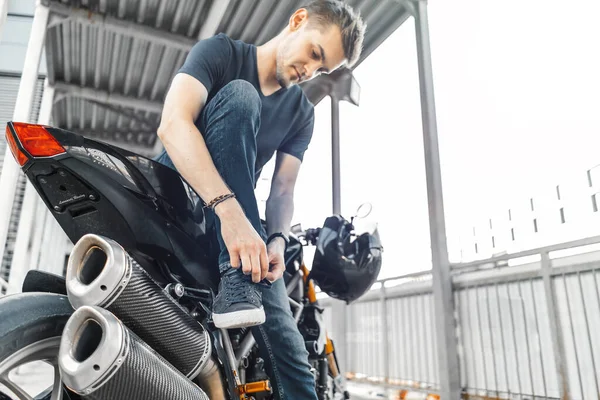 Motociclista guapo sentado en la motocicleta y enlazando zapatos en el aparcamiento . —  Fotos de Stock