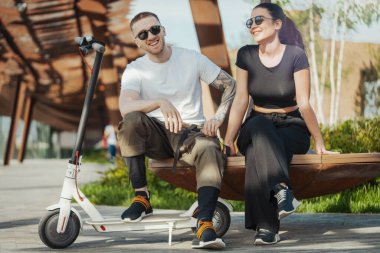 Modern park manzarasında elektrikli scooter 'ın yanında oturan ve gülen bir çift..