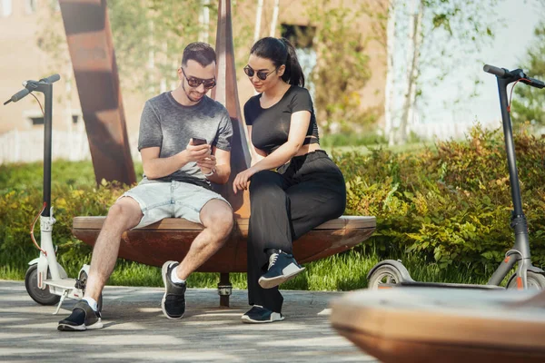 Pár sedí vedle elektrický koloběžka v moderní krajině parku a při pohledu na mobilní telefon. — Stock fotografie