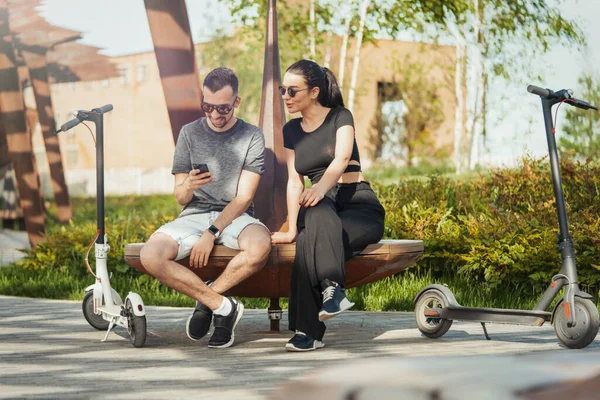 Pareja sentada al lado del scooter eléctrico en el paisaje moderno del parque y mirando el teléfono móvil . —  Fotos de Stock