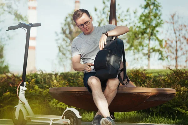 Attractive man talking by mobile phone and sitting next to electric kick scooter at modern park landscape. — Stok Foto