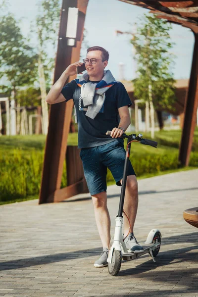 Attraktiv man talar med mobiltelefon och står bredvid elektrisk sparkcykel i modern park landskap. — Stockfoto