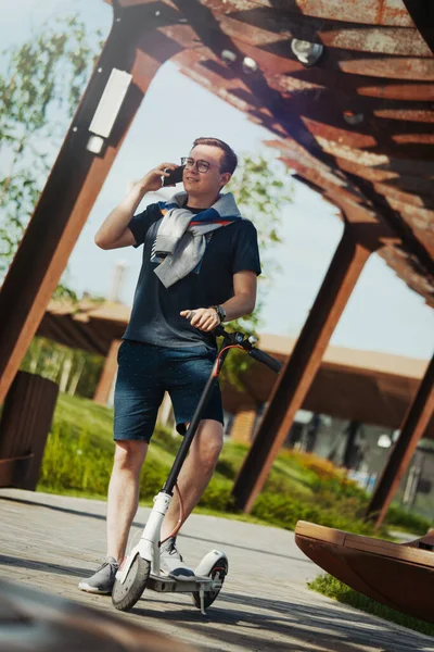 Attraktiv man talar med mobiltelefon och står bredvid elektrisk sparkcykel i modern park landskap. — Stockfoto