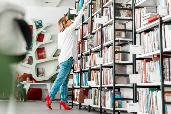 Young attractive pensive woman drawing hand to a book in a book store or library. Woman is on focus. Shelf with books are on foreground and blurred.