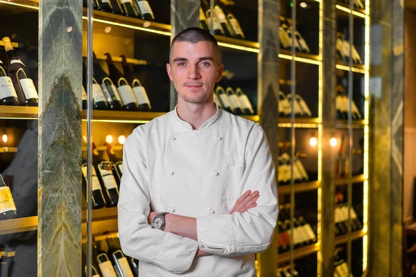 Young handsome cook chief posing in modern restaurant interior at wine refrigerator background.