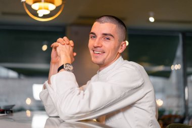 Beyaz üniformalı, modern lüks restoran içinde poz veren yakışıklı aşçı şefi. Esmerleştirilmiş arkaplanlı fotoğraf