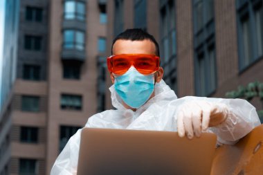 COVID Koruyucu Giysisi, Maskesi ve Gözlüğü olan ve Gökdelen Yakınlarındaki Güzel Şehir Manzarası 'nda laptopta çalışan yakışıklı serbest çalışan adam.