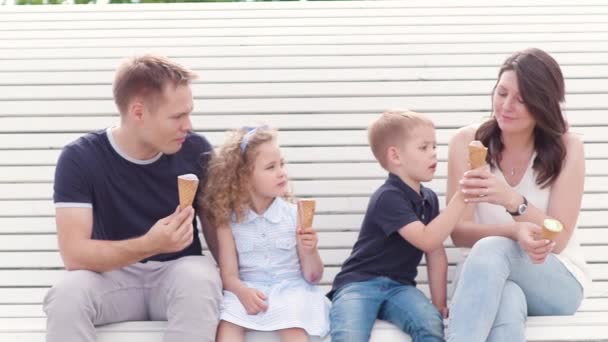 Familie-moeder, vader, zoon en dochter zitten op de Bank in de zomer vertellen verhalen lachen en eten van ijs in slow motion — Stockvideo