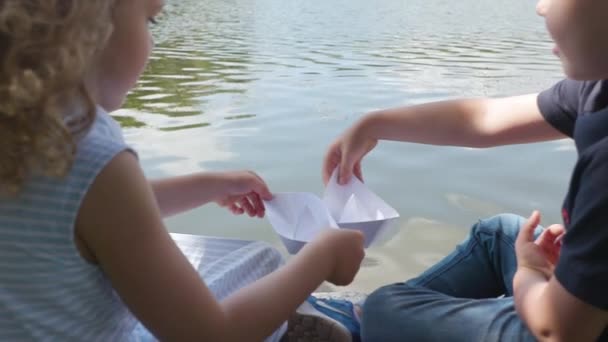 Fiú és lány játszani egy papír csónak, közel a folyó lassú mozgás — Stock videók