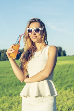 Kıvırcık saçlı gözlüklü genç bir ince kız gülümsüyor ve güneşli yaz gün gülümseyerek üzerinde bir şişe kamışla bir alkollü veya alkolsüz kokteyl içer