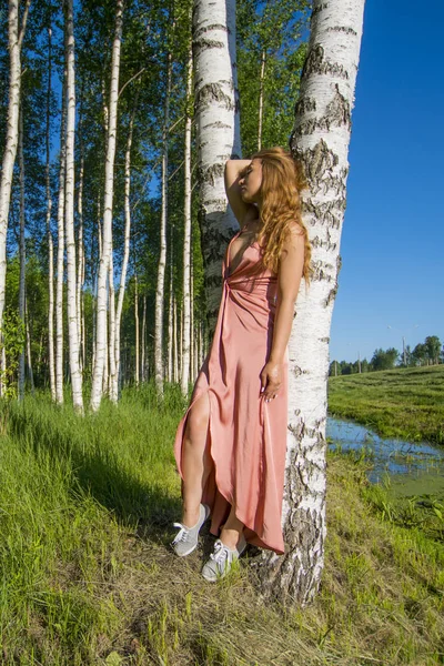 Joven esbelta hermosa chica en un vestido de coral largo posando en un bosque de abedul sonriendo de la mano cerca de la cabeza — Foto de Stock