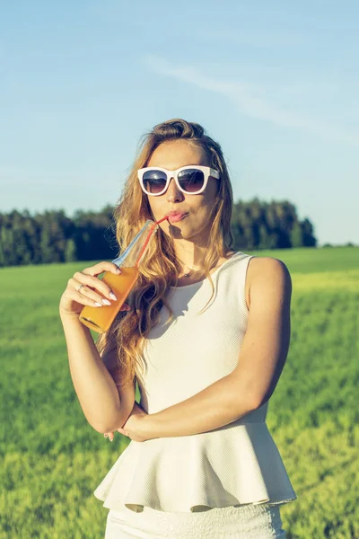 Kıvırcık saçlı gözlüklü genç bir ince kız gülümsüyor ve alkollü veya alkolsüz kokteyl kamışla bir güneşli yaz gününde bir şişeden içer