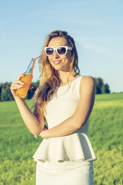 Kıvırcık saçlı gözlüklü genç bir ince kız gülümsüyor ve güneşli yaz gün gülümseyerek üzerinde bir şişe kamışla bir alkollü veya alkolsüz kokteyl içer