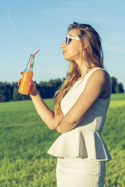 Kıvırcık saçlı gözlüklü genç bir ince kız gülümsüyor ve turuncu bir alkollü veya alkolsüz kokteyl kamışla bir şişeden bir güneşli yaz gününde içiyor.