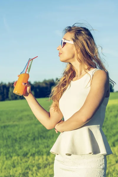 Kıvırcık saçlı gözlüklü genç bir ince kız gülümsüyor ve alkollü veya alkolsüz kokteyl kamışla dışında bir güneşli yaz gününde bir şişeden içer