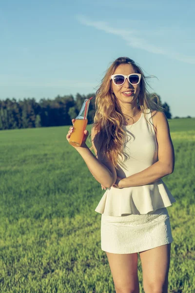 Kıvırcık saçlı gözlüklü genç bir ince kız gülümsüyor ve bir şişe üzerinde bir güneşli yaz gün açık havada bir saman bir alkollü veya alkolsüz kokteyl içer