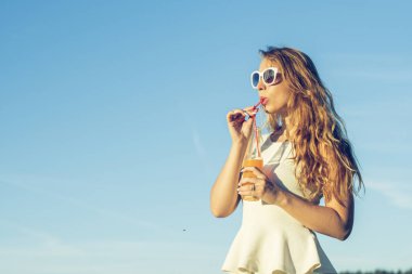 Kıvırcık saçlı gözlüklü genç bir ince kız gülümsüyor ve alkollü veya alkolsüz kokteyl kamışla bir güneşli yaz gününde bir şişeden içer