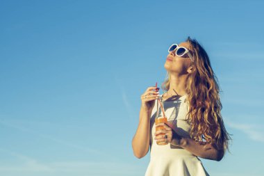 Kıvırcık saçlı gözlüklü genç bir ince kız gülümsüyor ve alkollü veya alkolsüz kokteyl kamışla bir güneşli yaz gününde bir şişeden içer