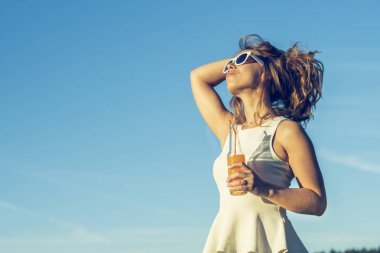 Kıvırcık saçlı gözlüklü genç bir ince kız gülümsüyor ve alkollü veya alkolsüz kokteyl kamışla saçını tutan bir güneşli yaz gününde bir şişeden içer