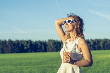 Kıvırcık saçlı gülümsüyor ve içecekler alkollü veya alkolsüz kokteyl bir şişe bir güneşli yaz gününde bir saman ve gülümseyerek gözlüklü genç bir ince kız