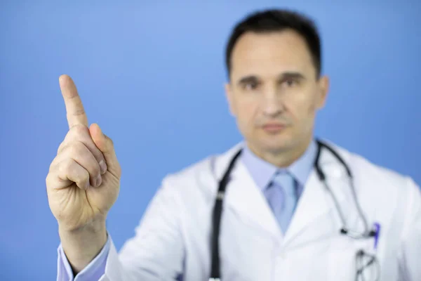 Serious doctor points up to empty text space on blue background, focus on finger — Stock Photo, Image