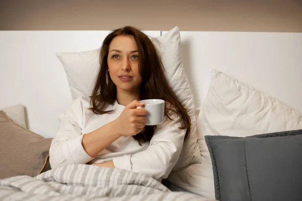 Νεαρή καυκάσια γυναίκα πίνει καφέ στο κρεβάτι και βλέπει την αγαπημένη του τηλεοπτική σειρά — Φωτογραφία Αρχείου
