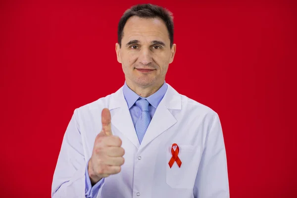 Älterer lächelnder Arzt im weißen Mantel mit roter Schleife darauf zeigt wie Zeichen — Stockfoto