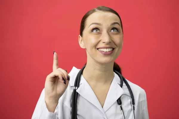 Emocionado atractivo médico apunta hacia arriba con el dedo al espacio de texto sobre fondo rojo —  Fotos de Stock