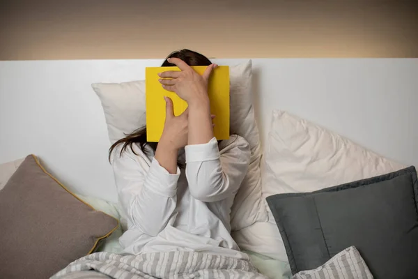 Καυκάσια κοπέλα με πιτζάμες στο κρεβάτι κρατά κίτρινο βιβλίο στα χέρια της και καλύπτει το πρόσωπό της — Φωτογραφία Αρχείου