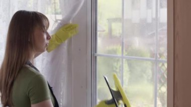 Ev hanımı cam temizleyicisi sıkar ve cam sileceği ile pencereyi siler.