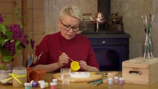Fokus artysta maluje powierzchnię żółtego garnka pędzlem na drewnianym stole. — Wideo stockowe
