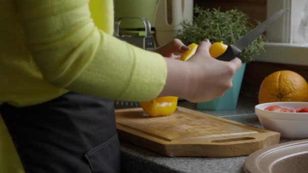 Ama de casa en delantal cortes amarillo pimentón dulce con cuchillo sobre tabla de madera — Vídeos de Stock