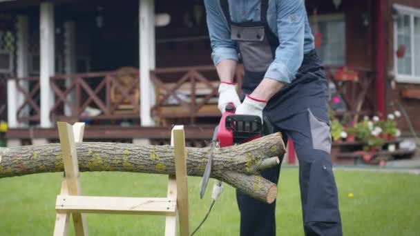 Man in workwear saws firewood on sawhorses with an electric saw at his home site — Stock Video