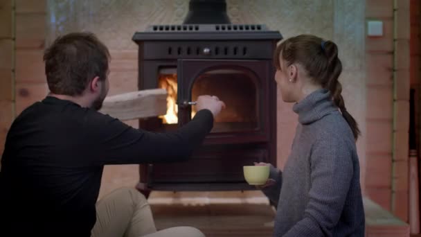 Schöne Mädchen trinkt Tasse Tee, Mann legt Brennholz in Kamin Paar ruht — Stockvideo