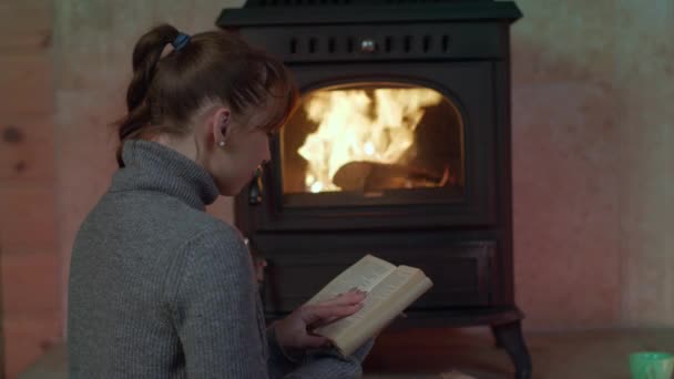 Güzel, genç bir kadın şöminenin yanında kâğıt kitap okuyor. — Stok video