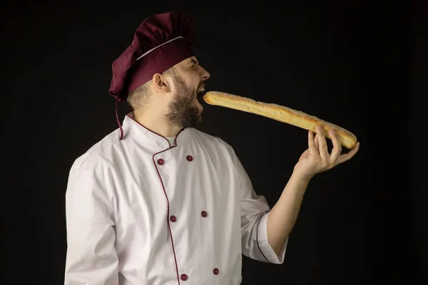 Handsome bearded chef man in uniform bites baguette, male baker cook